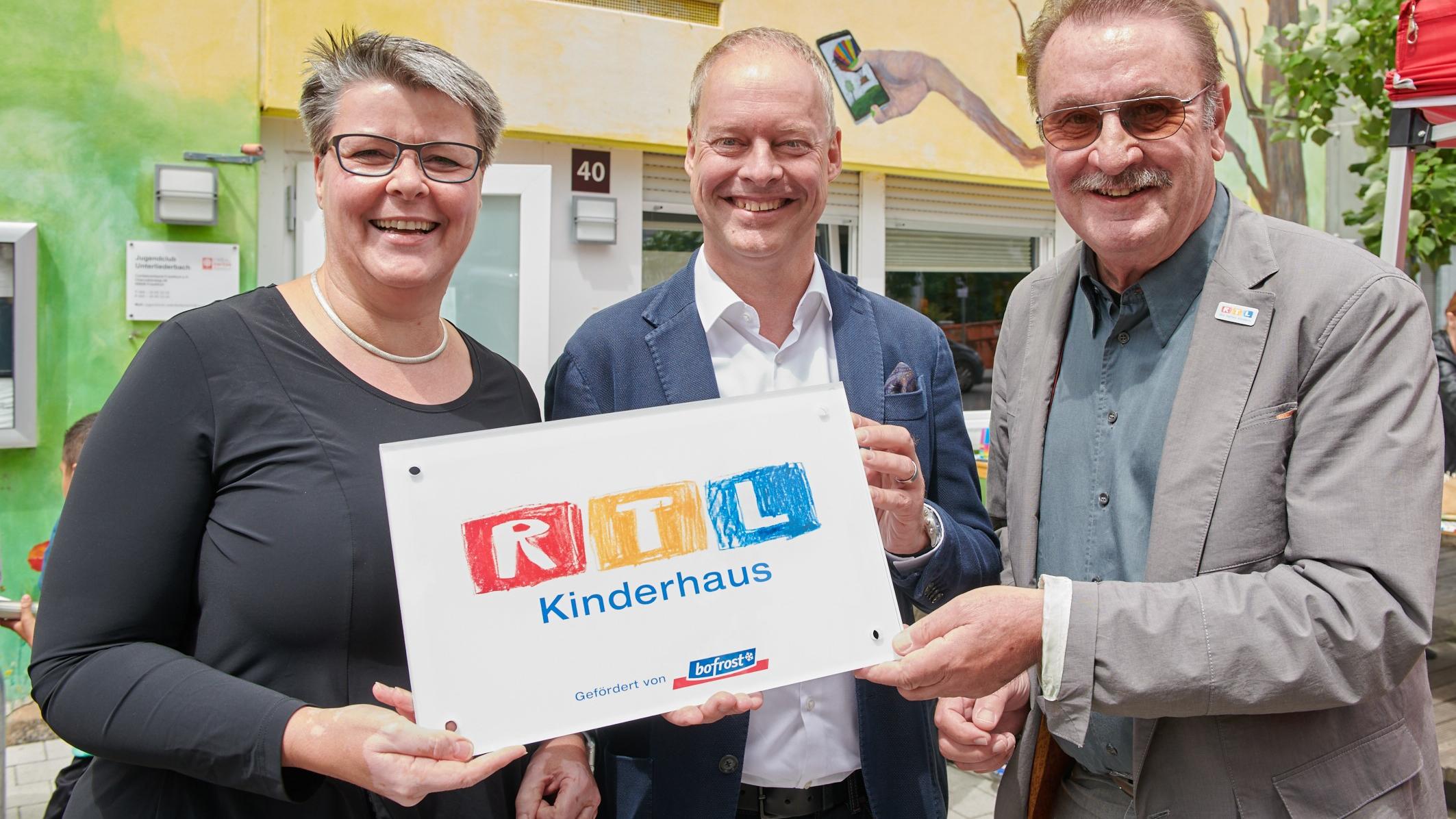 RTL-Kinderhaus Frankfurt: Zum 20. Jubiläum wird der Jugendclub Unterliederbach zum Kinderhaus Caritasdirektorin Gaby Hagmans, Dr. Axel Drösser, bofrost* Country Manager Deutschland und Achim Tirocke, Vorstand der „Stiftung RTL – Wir helfen Kindern e. V.“. mit dem Schild RTL-Kinderhaus