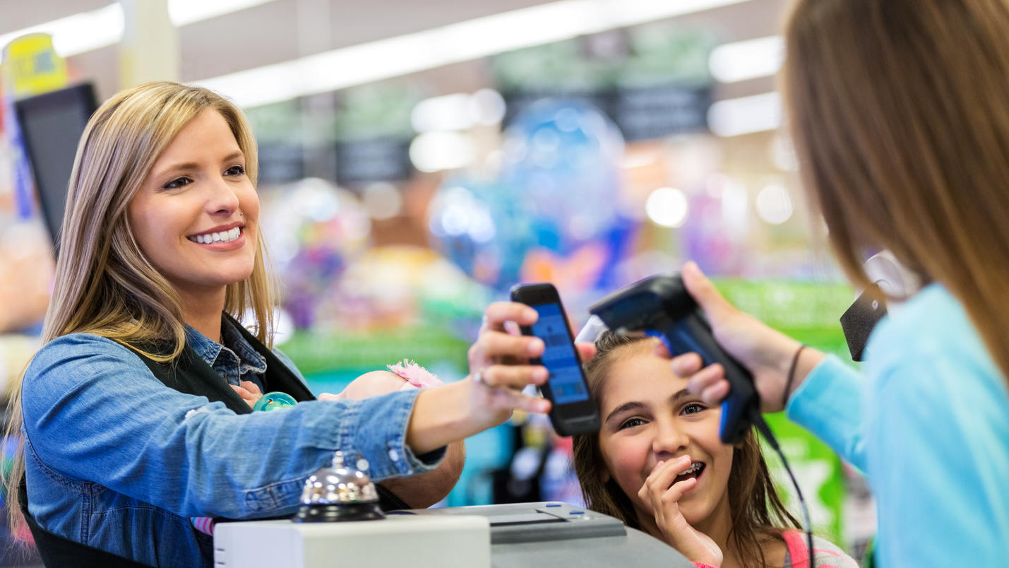 Kostenloses Geldabheben im Supermarkt: Die Supermarktkasse wird zum Geldautomat Mutter zeigt an der Supermarktkasse ihr Handy vor. Die Kassiererin scannt den Barcode auf dem Display.