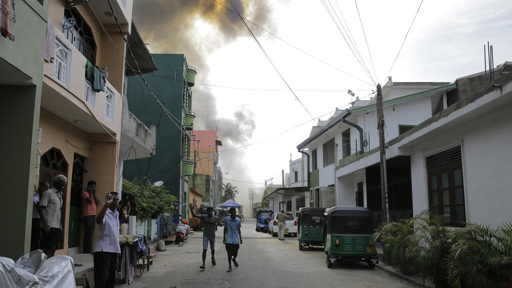 22.04.2019, Sri Lanka, Colombo: Menschen gehen auf einer Straße, während im Hintergrund Rauch aufsteigt. Am Ostermontag fand die Polizei weitere Sprengsätze in einem in der Nähe gesparkten Fahrzeug. Foto: Eranga Jayawardena/AP/dpa +++ dpa-Bildfunk ++