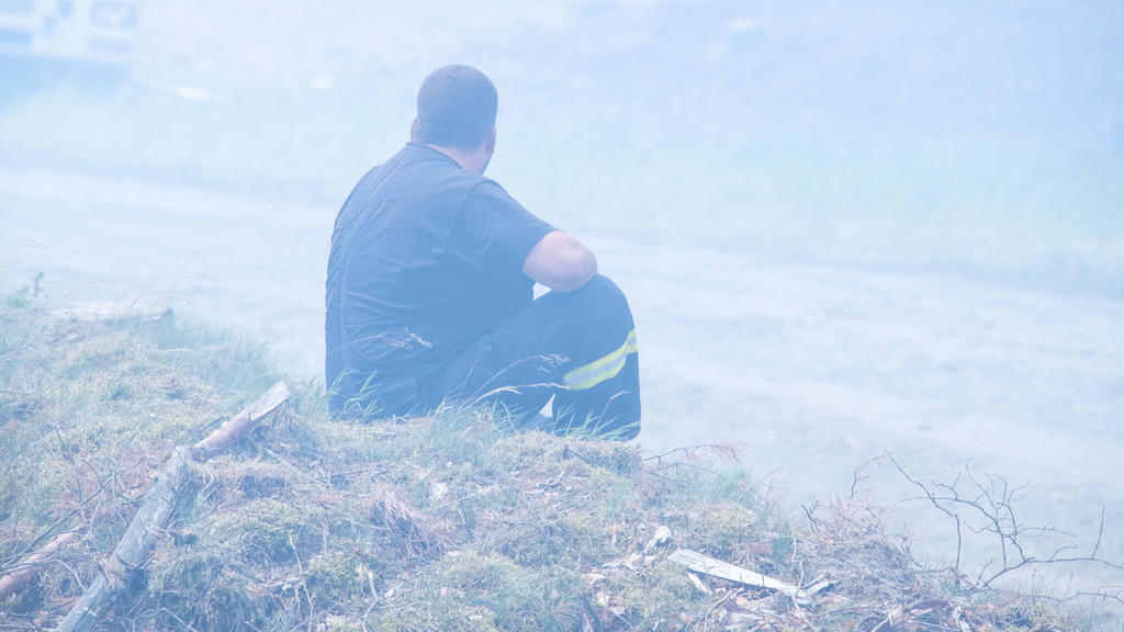 01.07.2019, Mecklenburg-Vorpommern, Alt Jabel: Ein erschöpfter Feuerwehrmann sitzt in einer Rauchwolke im brennenden Wald in der Nähe der kleinen Ortschaft Alt Kabel am Rande des ehemaligen Truppenübungsplatzes bei einem großflächigen Waldbrand in de
