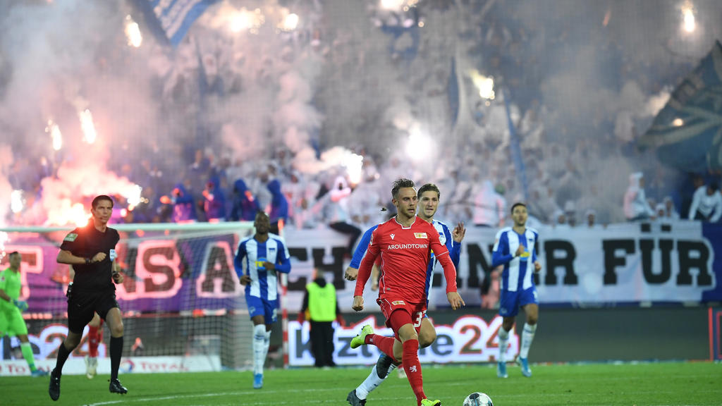 02.11.2019, Berlin: Fußball: Bundesliga, 10. Spieltag: 1. FC Union Berlin - Hertha BSC in der Alten Försterei. Unions Neven Subotic (M) führt den Ball während im Hintergrund Hertha Fans Pyrotechnik zünden. Links läuft Schiesrichter Deniz Aytekin, der
