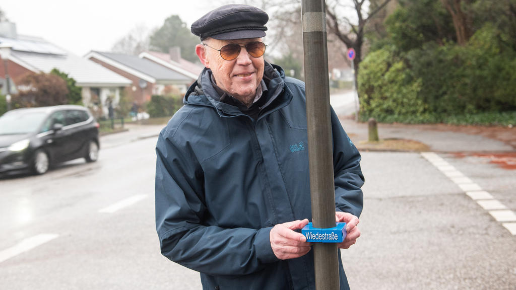 06.12.2019, Schleswig-Holstein, Wedel: Volker König, Erfinder von Hilfsmitteln für Behinderte, steht an einem Straßenschild, an dessen Mast ein zusätzliches kleines Schild für Sehbehinderte hängt. Die Stadt Wedel in Schleswig-Holstein will als erste