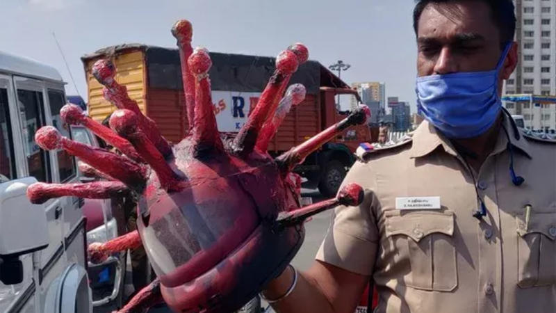 Der Polizist trägt einen Mundschutz und hält in der Hand einen roten Helm, der wie das Corona-Virus aussieht.