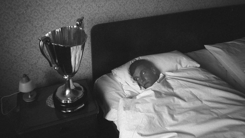 Franz Bulle ROTH liegt im Bett und schaut auf den Europapokal, der auf dem Nachttisch steht,