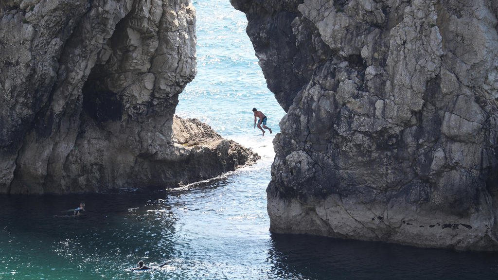 31.05.2020, Großbritannien, Lulworth: Ein Mann springt von der Durdle Door, einer natürlichen Felsbrücke aus Kalkstein, ins Meer, obwohl der Gemeinderat von Dorset den Strand für die Öffentlichkeit geschlossen hat, nachdem sich drei Menschen bei Sprü