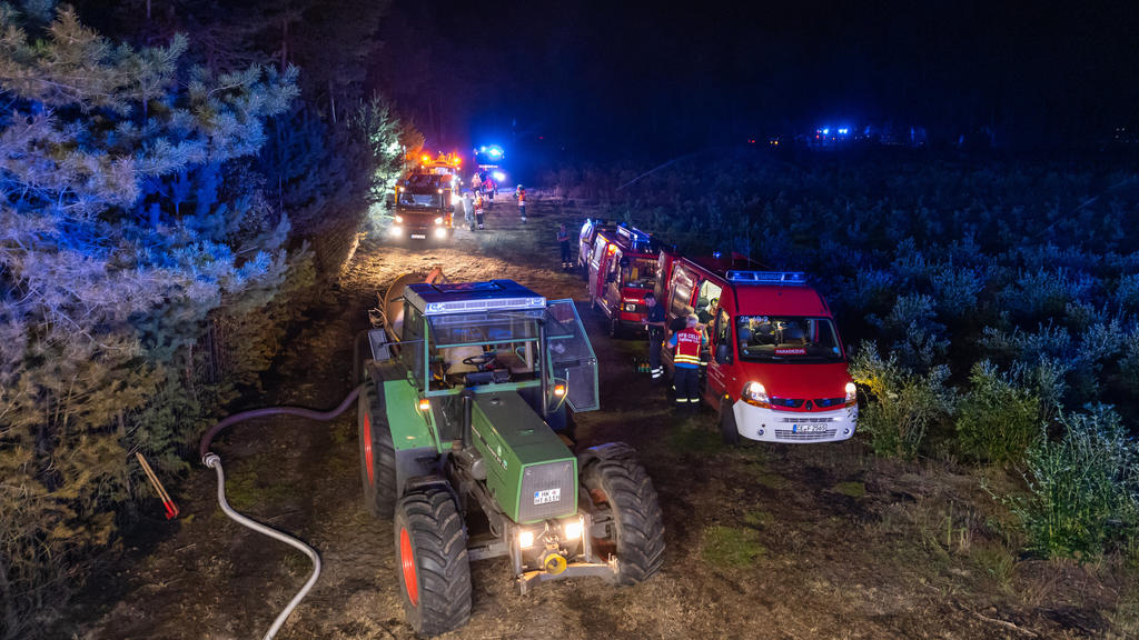 12.08.2020, Niedersachsen, Essel: Ein Trekker und Einsatzfahrzeuge der Feuerwehr stehen für Nachlöscharbeiten an einem Waldstück. Wegen eines großflächigen Brandes im Heidekreis ist das Wochenendgebiet Esseler Wald am Mittwochabend nach Polizeiangabe