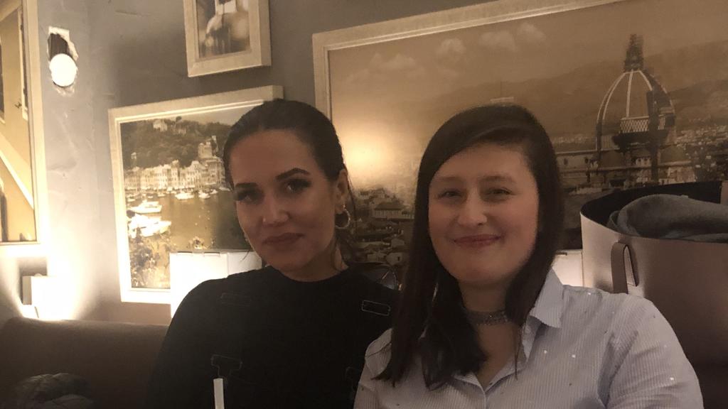 Zwei junge Frauen sitzen in einem Restaurant an einem Tisch und schaen freundlich in die Kamera.