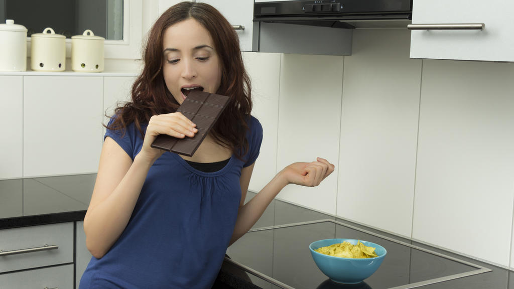 Junge Frau steht in der Küche und beißt in eine Tafel Bitterschokolade. Auf der Küchenzeile steht eine Schüssel mit Kartoffelchips.