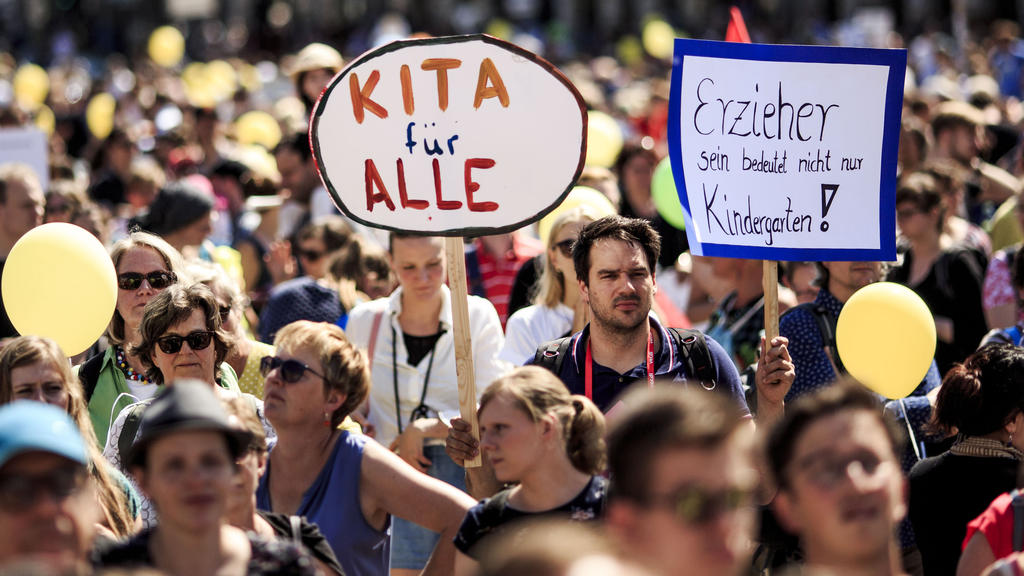 26.05.2018, Berlin: Eltern und Erzieher demonstrieren u.a. mit einem Schild "Kita für alle" für mehr Plätze in den Kitas und eine bessere Bezahlung der Erzieher. In Berlin gibt es laut Familienverwaltung rund 170 000 Plätze in Kitas und Tagespflege.